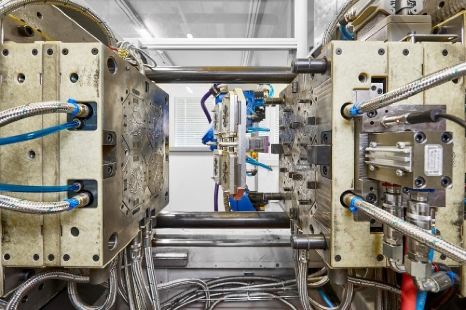 Nahaufnahme einer industriellen Spritzgießmaschine mit offenen Metallformen, sichtbaren Schläuchen, Kabeln und Ausrüstungskomponenten in einer sauberen, modernen Produktionsstätte.