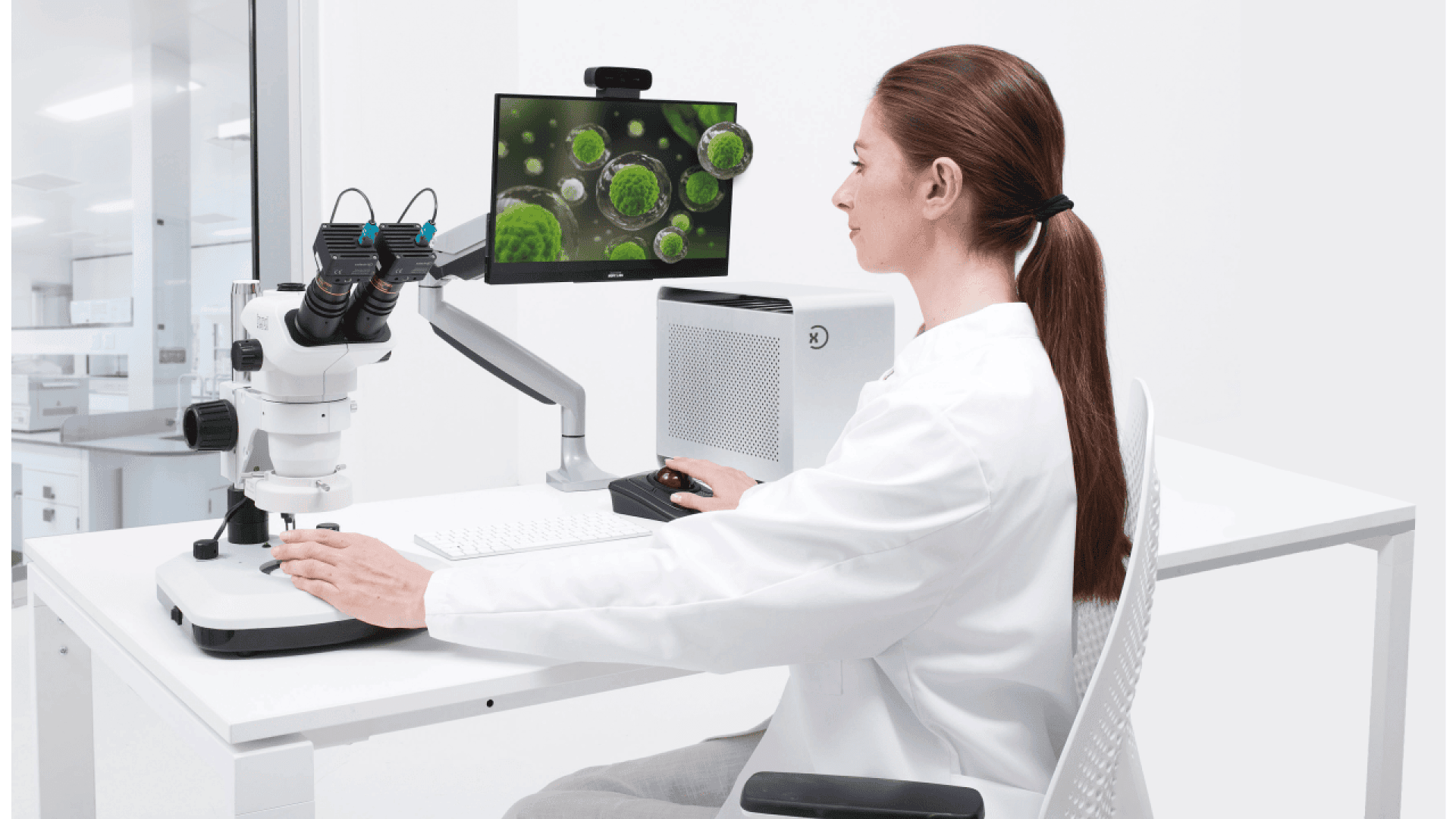A scientist in a white lab coat examines microorganisms on a monitor connected to a microscope in a modern laboratory setting. Laboratory equipment and a computer are visible on the white desk.