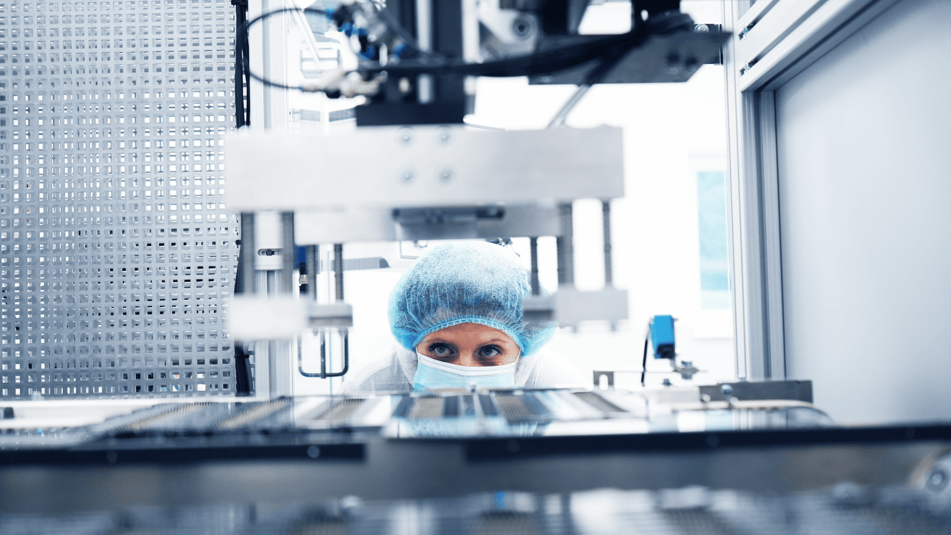 Eine Person mit blauem Haarnetz, Maske und Laborkittel beobachtet Industriemaschinen in einem modernen, sterilen Labor oder einer Produktionsanlage.