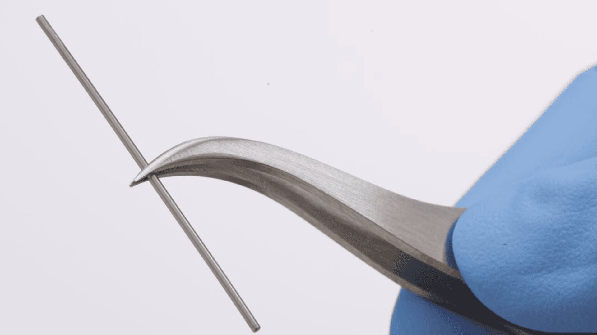 A gloved hand holds a curved metal tweezer gripping a thin, straight metal rod against a plain, light background.
