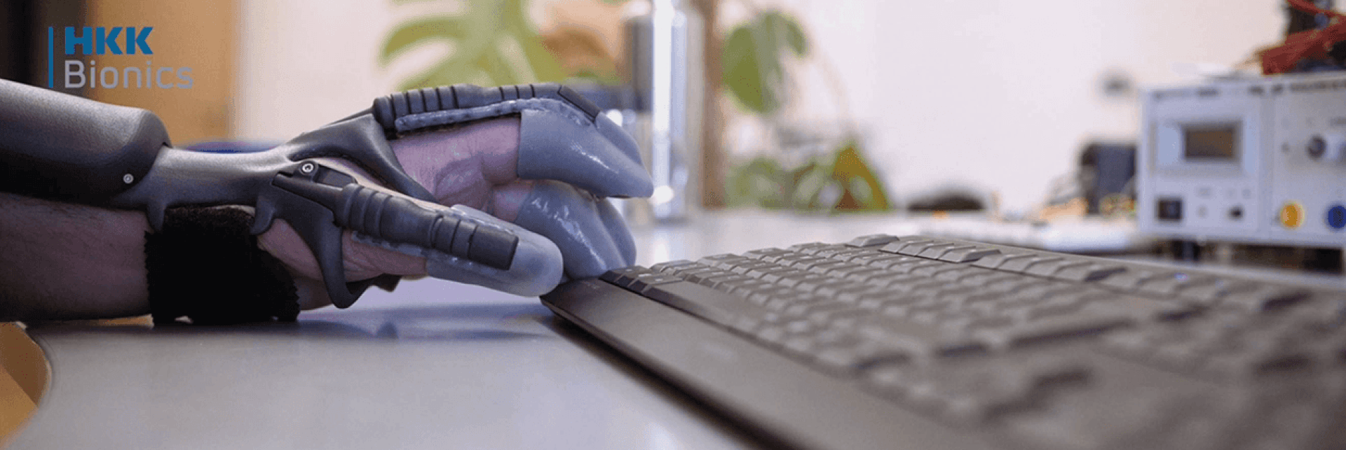 A person wearing a gray robotic hand prosthesis is typing on a black computer keyboard. The HKK Bionics logo is visible in the upper left corner, and electronic devices are on the desk.