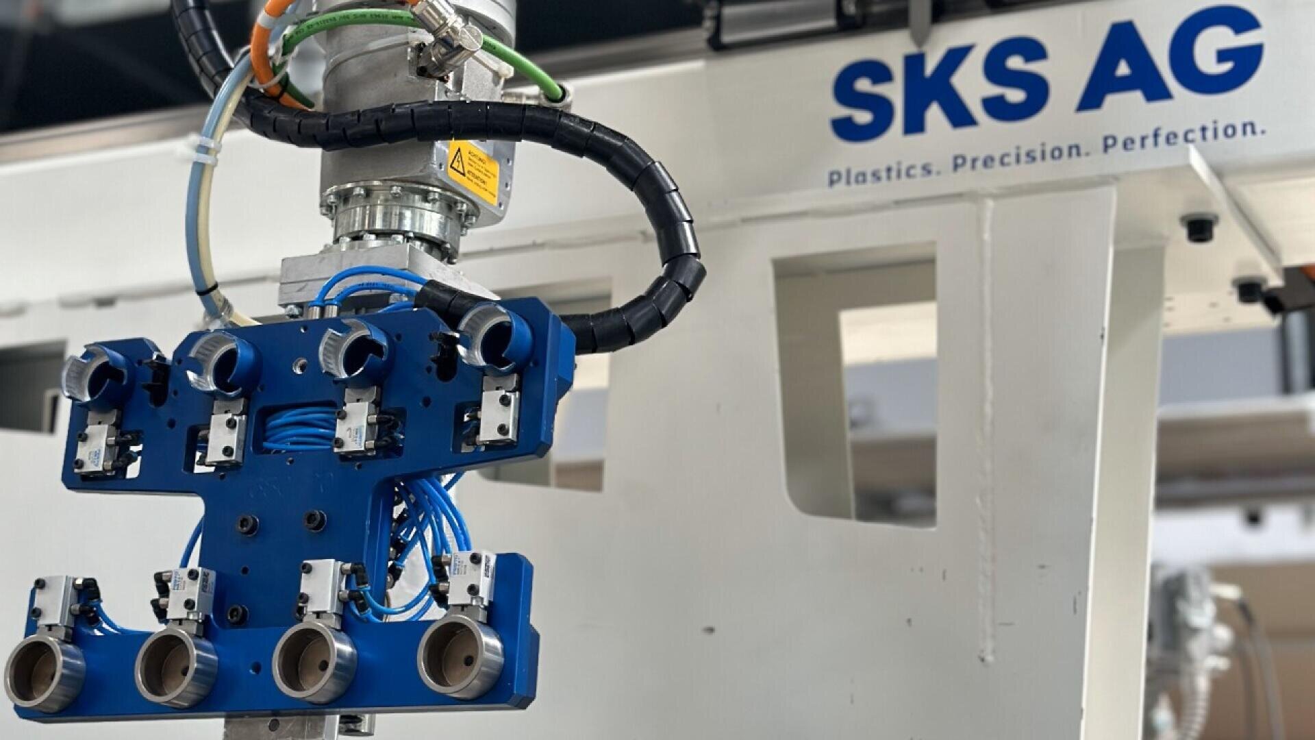 A close-up of a robotic arm with blue mechanical grippers and wires, attached to industrial equipment labeled SKS AG Plastics. Precision. Perfection. in blue text.