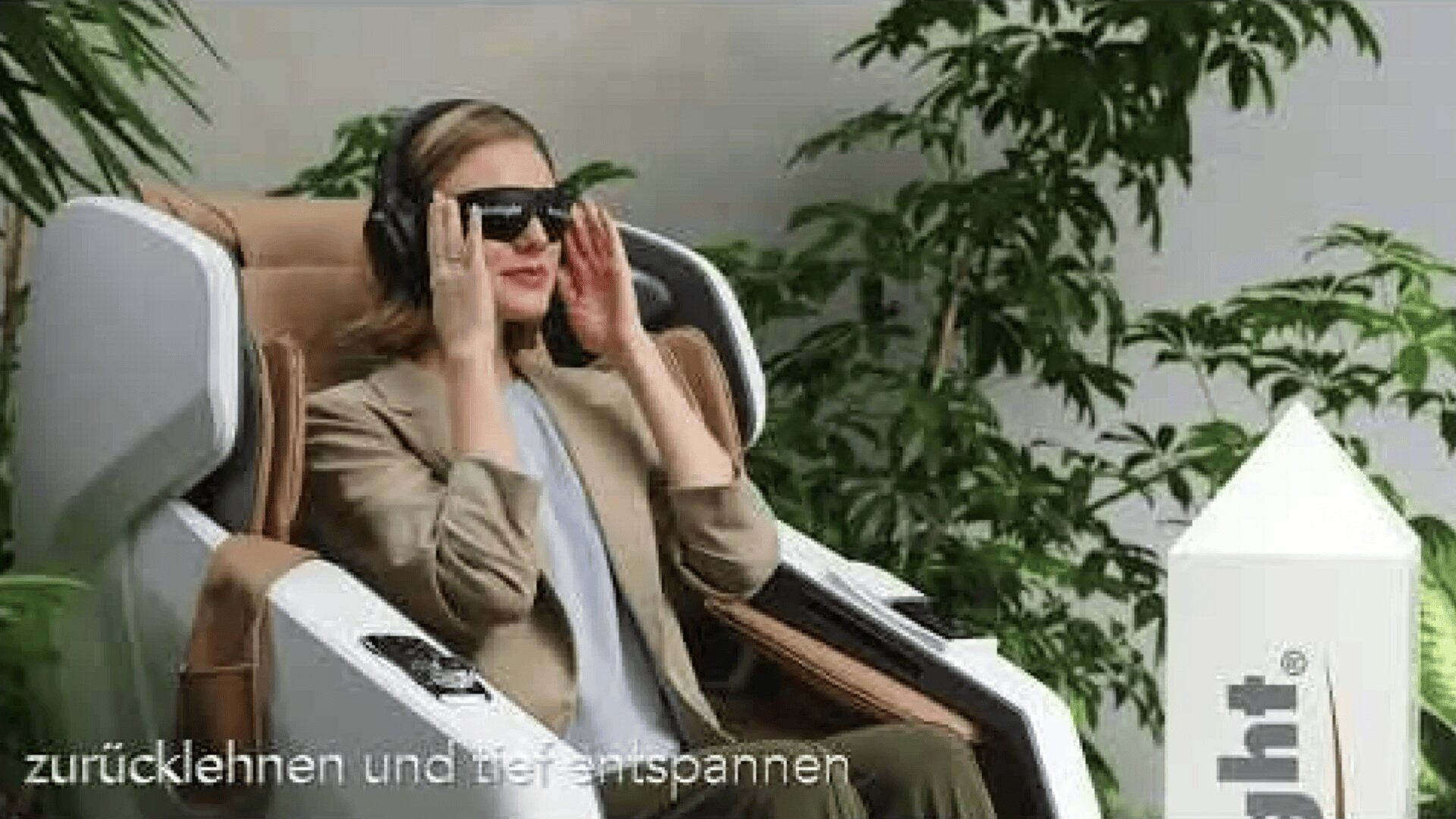 Eine Frau sitzt in einem modernen Massagestuhl. Sie trägt Kopfhörer und eine dunkle Brille, lächelt und entspannt sich. Im Hintergrund sind grüne Pflanzen zu sehen, darunter ein deutscher Text.