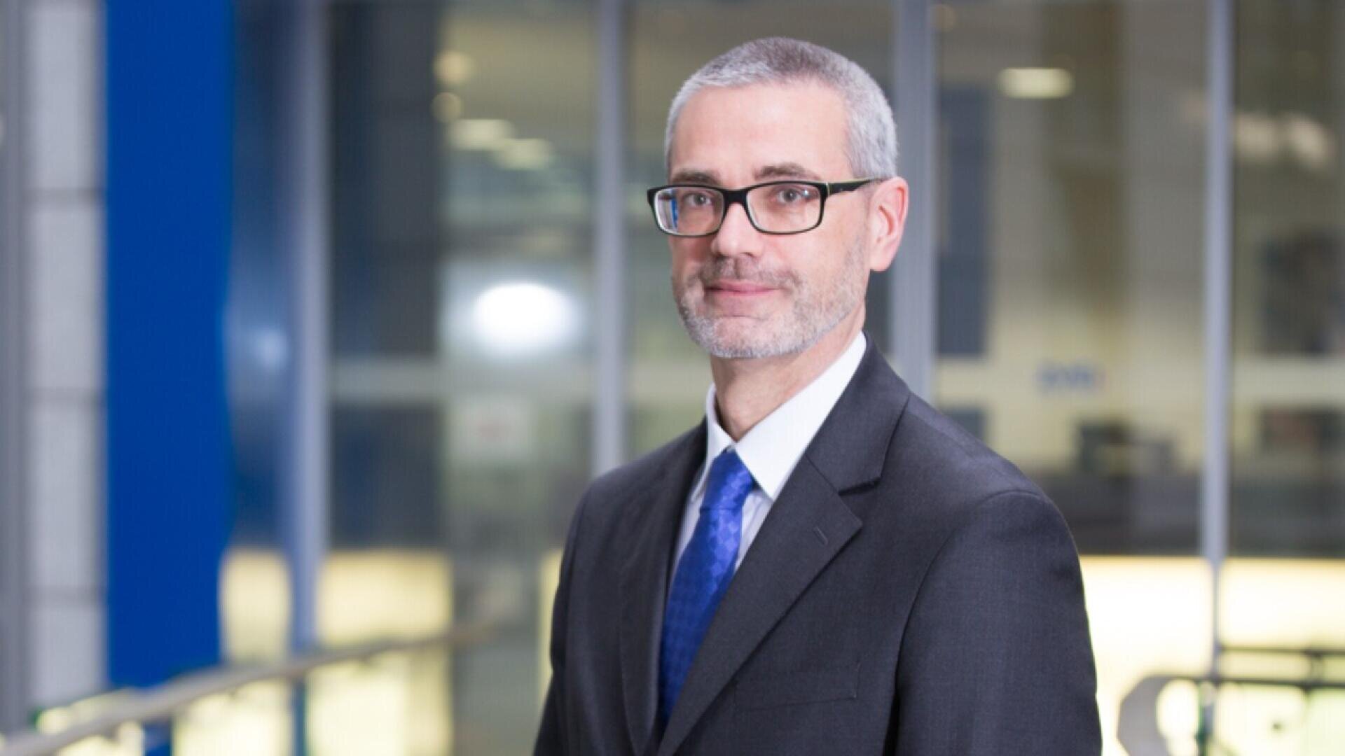 A man with short gray hair, glasses, and a trimmed beard is wearing a dark suit, white shirt, and blue tie, standing indoors in front of large windows with blurred office background.