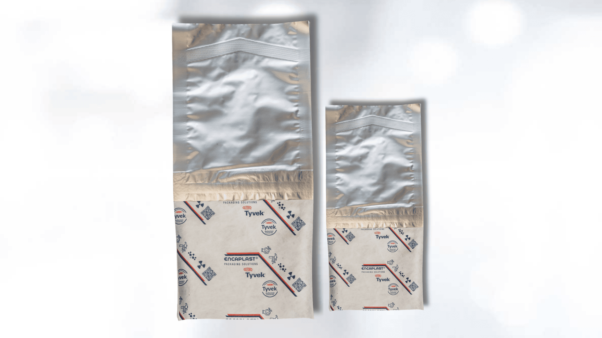 Two silver and white medical sterilization pouches with a blue and red printed Tyvek part, shown side by side on a light, blurred background.