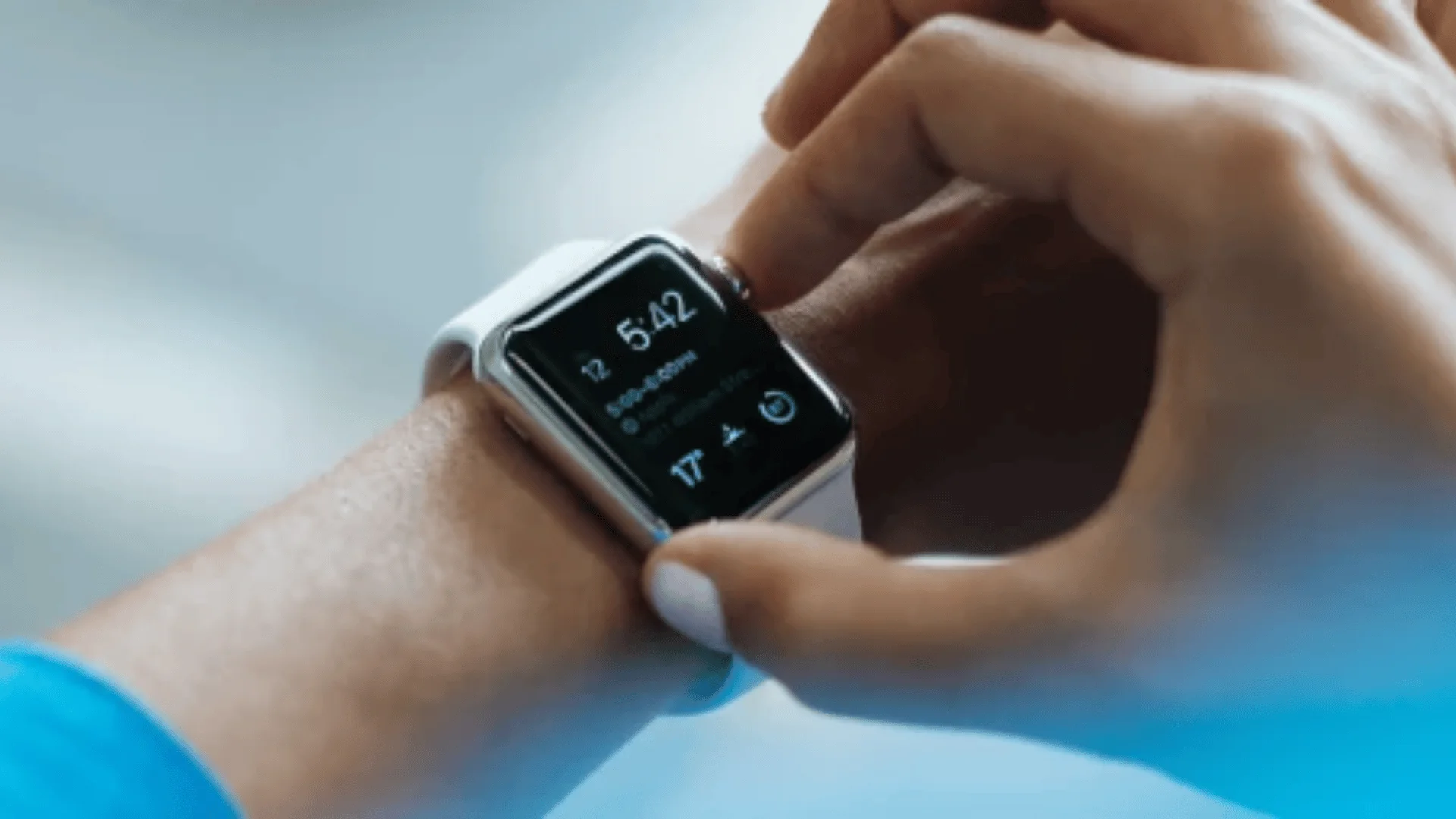 Eine Person interagiert mit einer Smartwatch an ihrem Handgelenk und berührt den Bildschirm, der Uhrzeit (5:42), Datum, Wetter und verschiedene Symbole anzeigt. Das Armband der Uhr ist weiß, der Hintergrund ist unscharf.