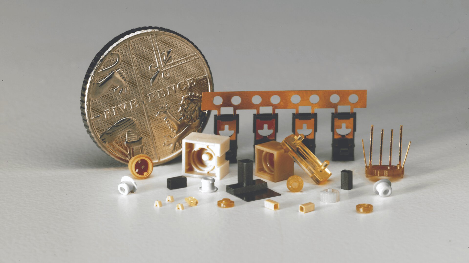 A British five pence coin stands upright next to a variety of tiny gold, white, and black electronic components on a white surface, highlighting their small size in comparison.