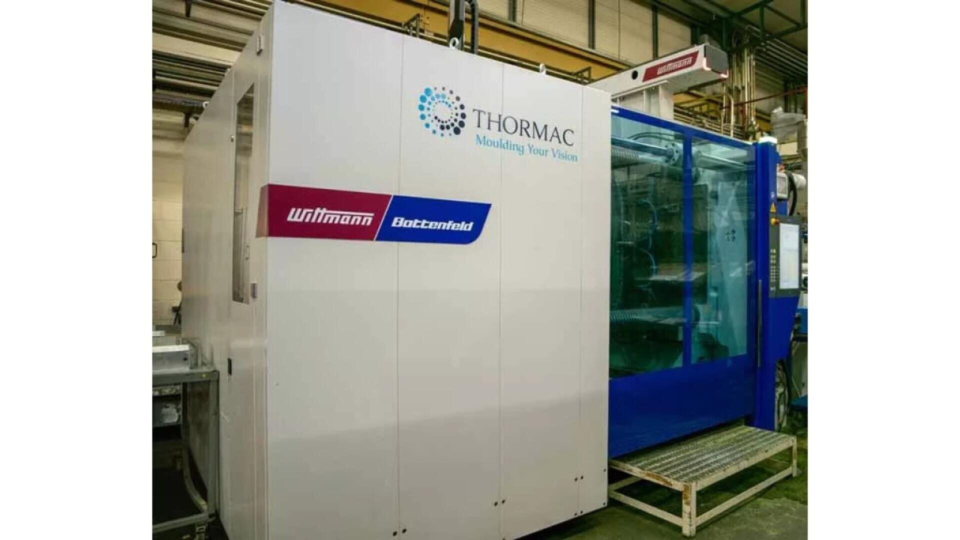 Eine große industrielle Spritzgussmaschine mit den Marken THORMAC, Wittmann und Battemfeld in einer Fabrikumgebung mit einem weiß-blauen Äußeren und durch eine durchsichtige Platte sichtbaren Innenkomponenten.