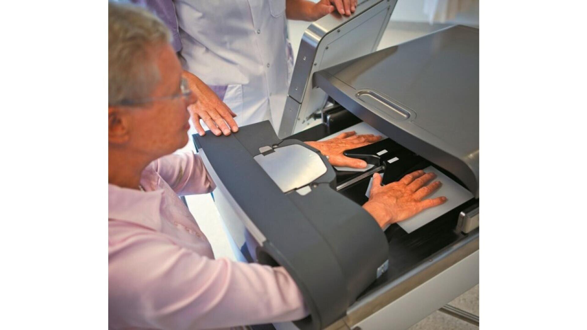 Eine ältere Person legt beide Hände auf einen Scanner, während ein medizinisches Fachpersonal assistiert und einen medizinischen oder Identifikationsscan in einer klinischen Umgebung vorschlägt.