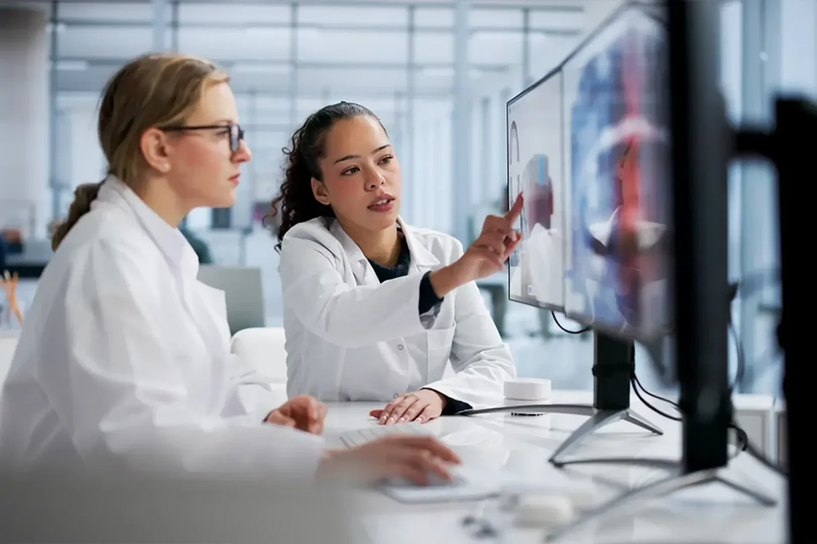 Zwei Wissenschaftler in weißen Laborkitteln sitzen an einem Schreibtisch und schauen auf Computermonitore. Der eine zeigt auf den Bildschirm, während der andere tippt. Beide konzentrieren sich auf ihre Arbeit in einem hellen, modernen Laborumfeld.