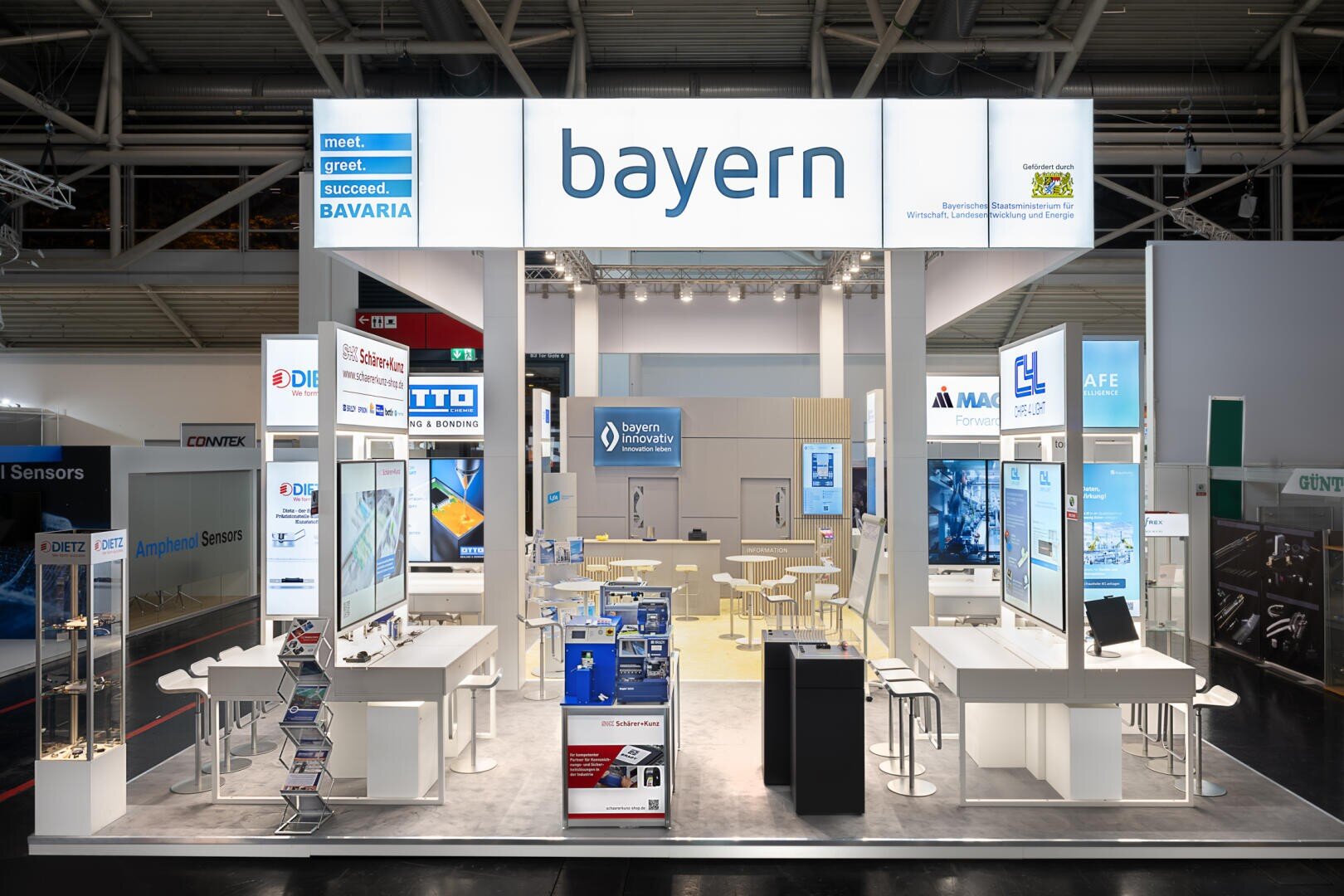 A brightly lit exhibition stand with a large bayern sign overhead, screens, counters, stools, brochures and banners presenting Bavaria's technology and innovation brands.