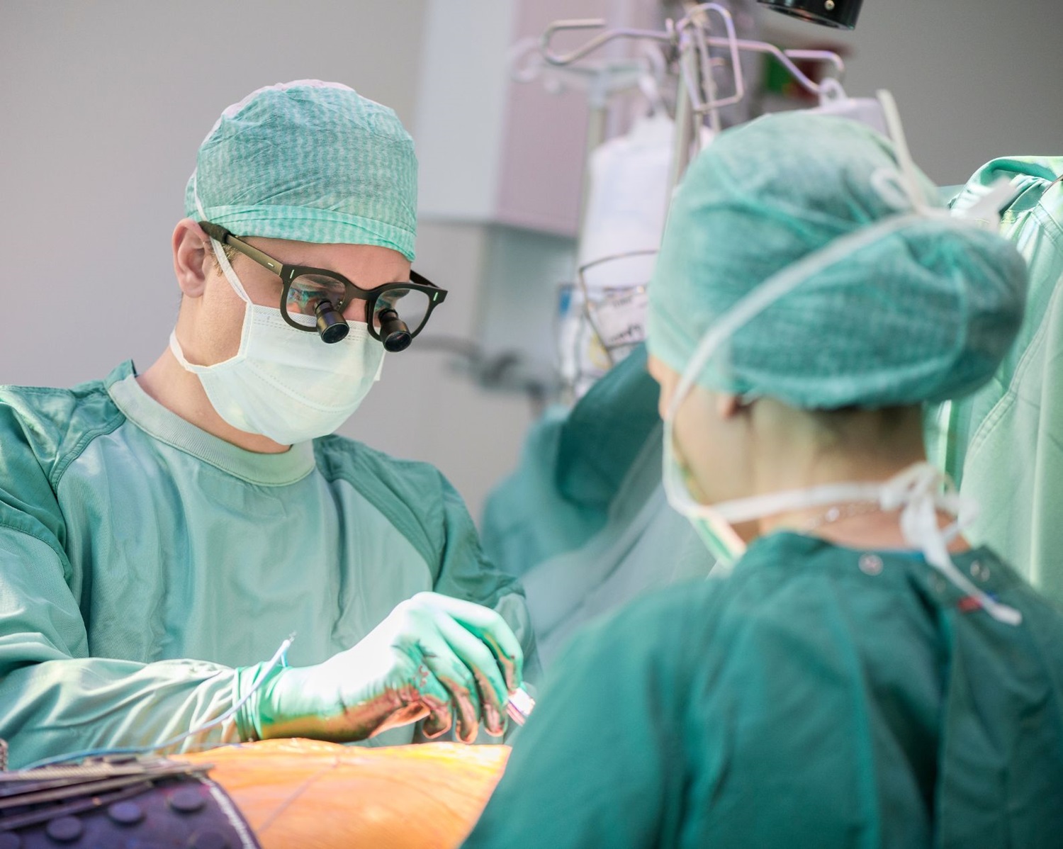 Zwei Chirurgen in grünen Kitteln, Masken und Mützen führen in einem Operationssaal eine Operation durch. Ein Chirurg trägt eine Lupenbrille und konzentriert sich auf den Eingriff, während der andere assistiert.