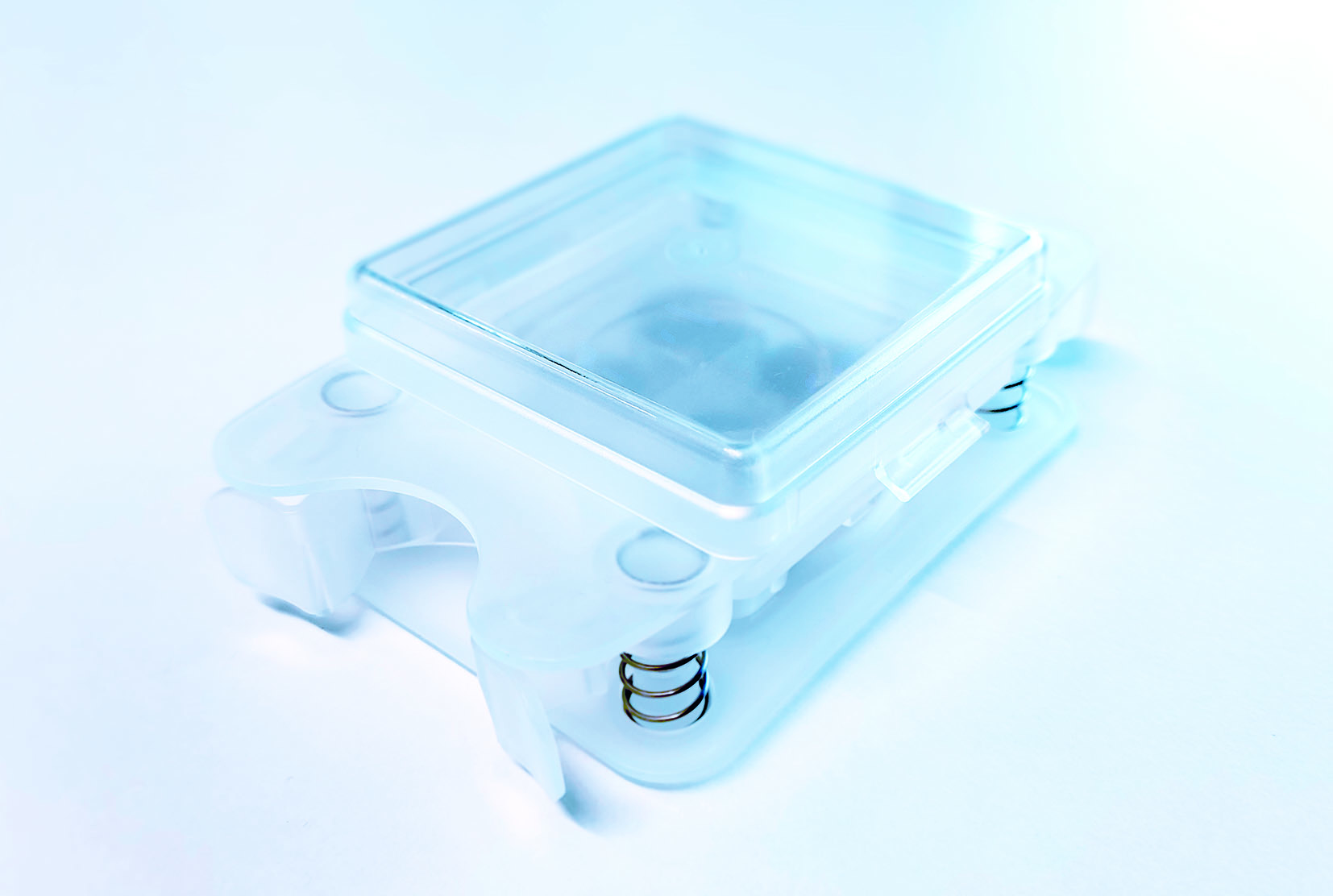 Nahaufnahme eines elektronischen Bauteils aus transparentem Kunststoff mit einem quadratischen Deckel, sichtbaren Metallfedern und weißem Hintergrund.