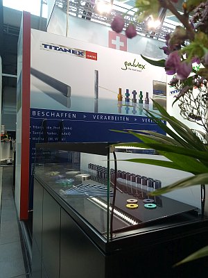 Auf einem Messestand werden elektrische Kabel und bunte Drahtspulen in einer Glasvitrine ausgestellt. Dar&uuml;ber befinden sich gro&szlig;e Schilder mit den Marken TITANEX und Galvex, und im Vordergrund sind teilweise gr&uuml;ne Pflanzen zu sehen.