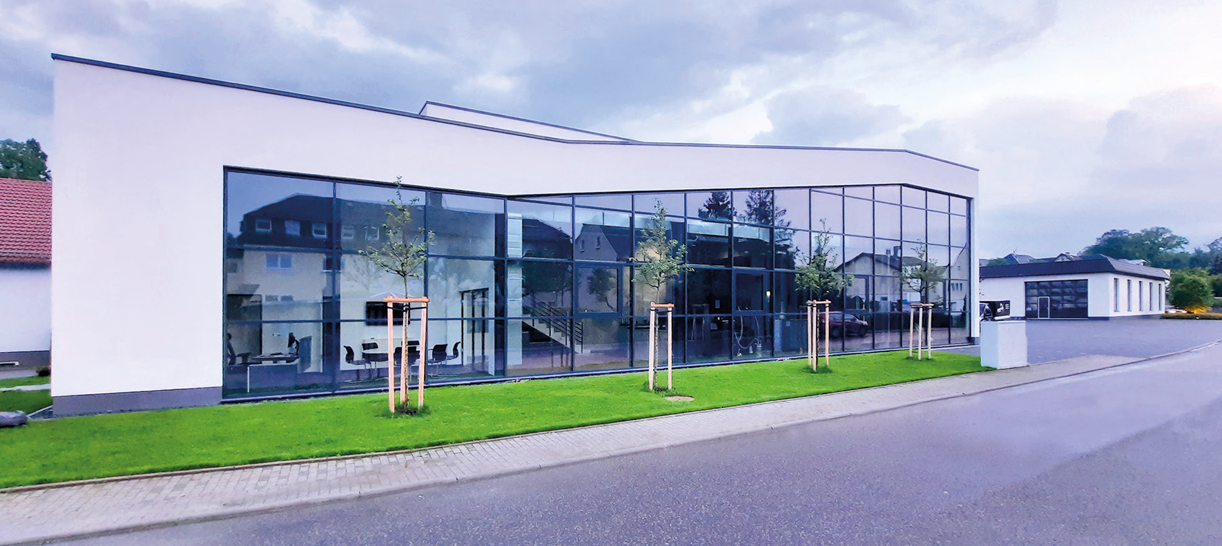 Modernes Gebäude mit großen Glasfenstern, weißen Außenwänden und einem Flachdach, umgeben von grünem Gras und jungen Bäumen, an einer gepflasterten Straße unter bewölktem Himmel gelegen.