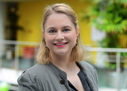 Eine Frau mit schulterlangen blonden Haaren, die einen grauen Blazer und ein schwarzes Oberteil trägt, lächelt in eine Kamera in einem hellen Innenraum mit grünen Pflanzen und einem gelben Hintergrund.