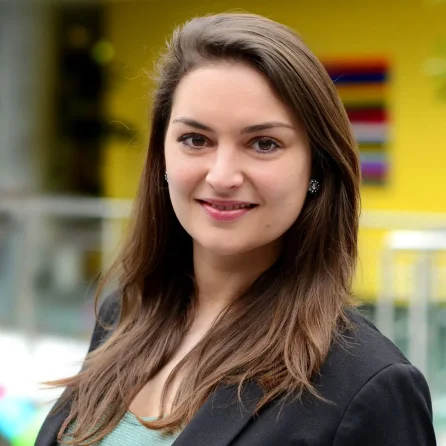 Eine Frau mit langen braunen Haaren lächelt in die Kamera. Sie trägt einen dunklen Blazer und ein helles Oberteil und steht vor einem leuchtend gelben Hintergrund mit bunten, verschwommenen Verzierungen.