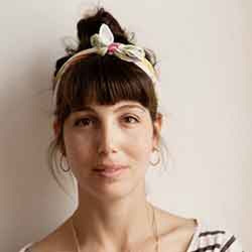 Eine Frau mit dunklen Haaren in einem Dutt, die ein geblümtes Stirnband und Reifohrringe trägt, blickt mit neutralem Blick in die Kamera. Sie trägt ein gestreiftes Hemd und eine Halskette und steht vor einem schlichten hellen Hintergrund.