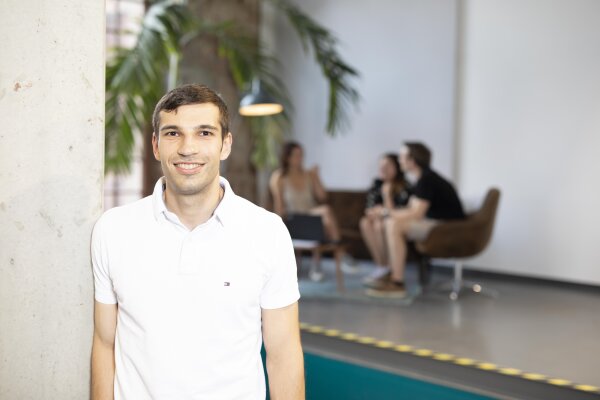A young man in a white polo shirt smiles at the camera in a modern office. In the background, three people sit on a sofa next to a potted plant, chatting.