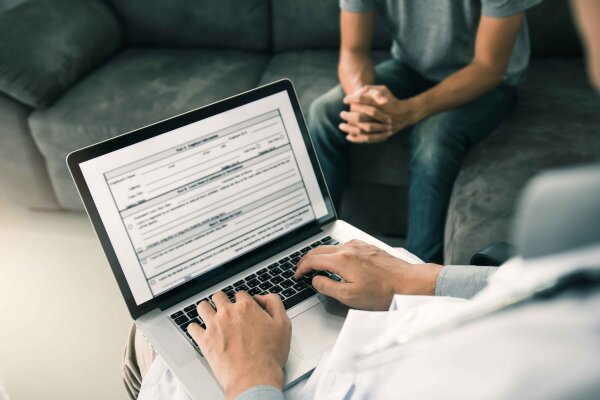 Ein Arzt tippt Notizen auf einem Laptop, während ein Patient in der Nähe auf einer Couch sitzt und die Hände verschränkt, was an eine Konsultation oder ein medizinisches Gespräch in einer klinischen Umgebung erinnert.