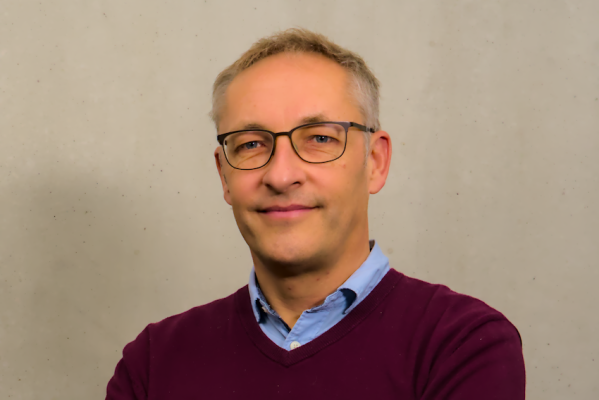 Ein Mann mittleren Alters mit kurzen grauen Haaren und Brille, der ein blaues Hemd mit Kragen unter einem kastanienbraunen Pullover trägt, steht vor einem einfarbig beigen Hintergrund, schaut in die Kamera und lächelt leicht.