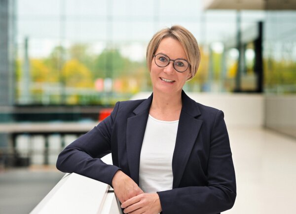 A woman with short blonde hair and glasses, wearing a navy blazer and white shirt, stands indoors and smiles at the camera. The background is modern, with large windows and soft natural light.