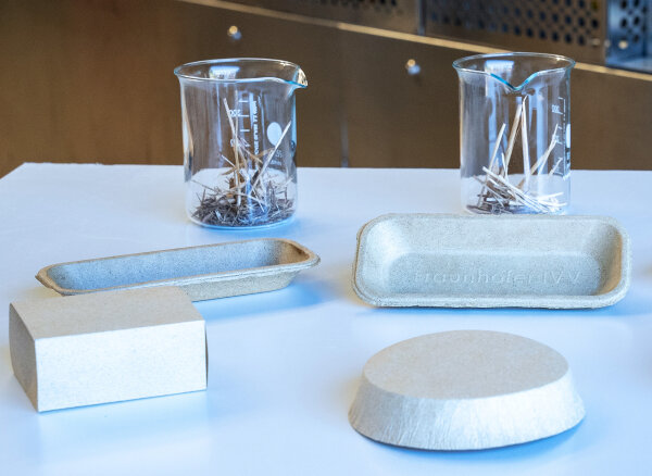 Two cups filled with small pieces of wood stand behind four prototypes of molded fiber packaging, including a tray, a rectangular block and a shallow bowl displayed on a white surface.