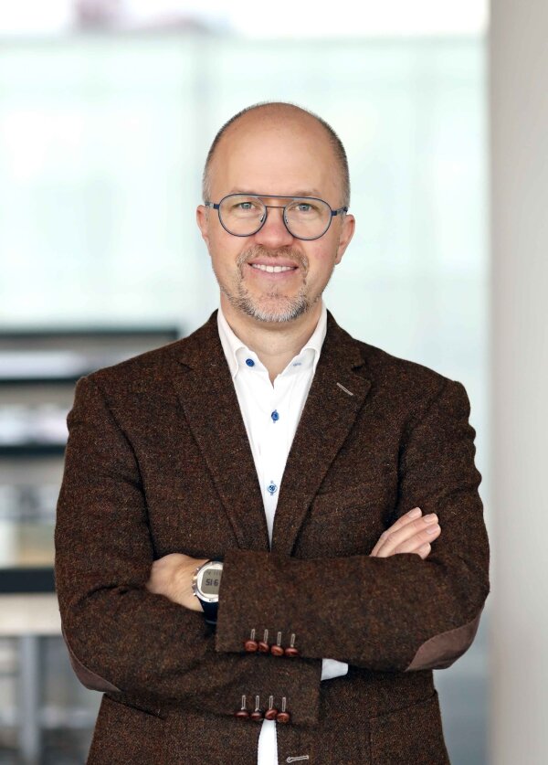 Ein lächelnder Mann mit Brille, Bart und braunem Blazer steht mit verschränkten Armen in einer hellen, modernen Inneneinrichtung.