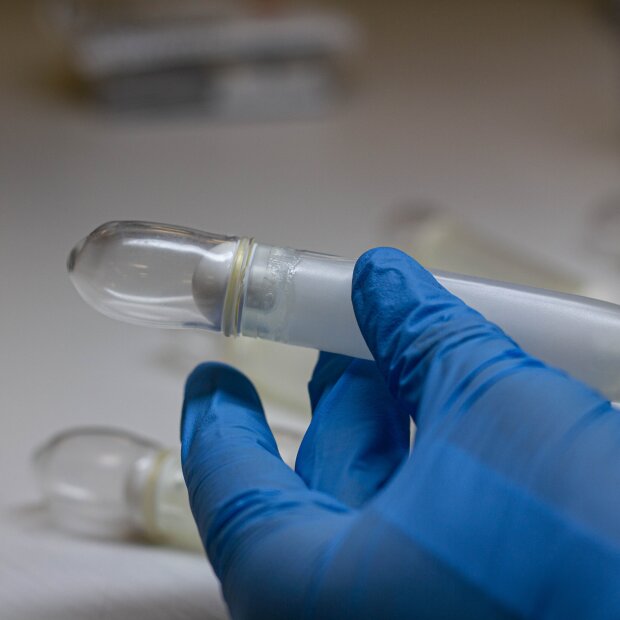 Eine Hand mit einem blauen Handschuh hält ein transparentes, kapselförmiges Laborröhrchen mit verschlossenem Ende. Andere ähnliche Röhrchen sind im Hintergrund auf einer weißen Fläche verschwommen zu erkennen.