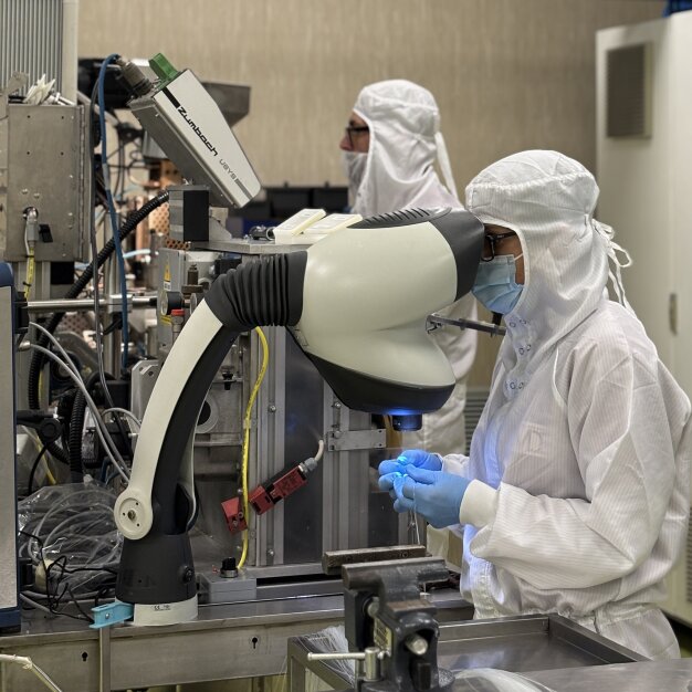 Zwei Personen in weißen Reinraumanzügen und Masken arbeiten in einem Labor mit modernen Geräten. Eine Person bedient ein großes mikroskopähnliches Gerät, während sie mit blauen Handschuhen ein kleines Objekt hält.
