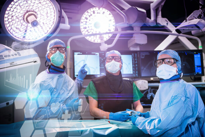 Three surgeons in gowns, masks and goggles operate in a high-tech operating theater with bright lights, monitors and digital medical graphics superimposed in the foreground.