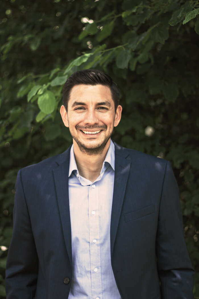A man in a dark blue suit jacket and a light blue shirt smiles as he stands outside in front of green foliage.