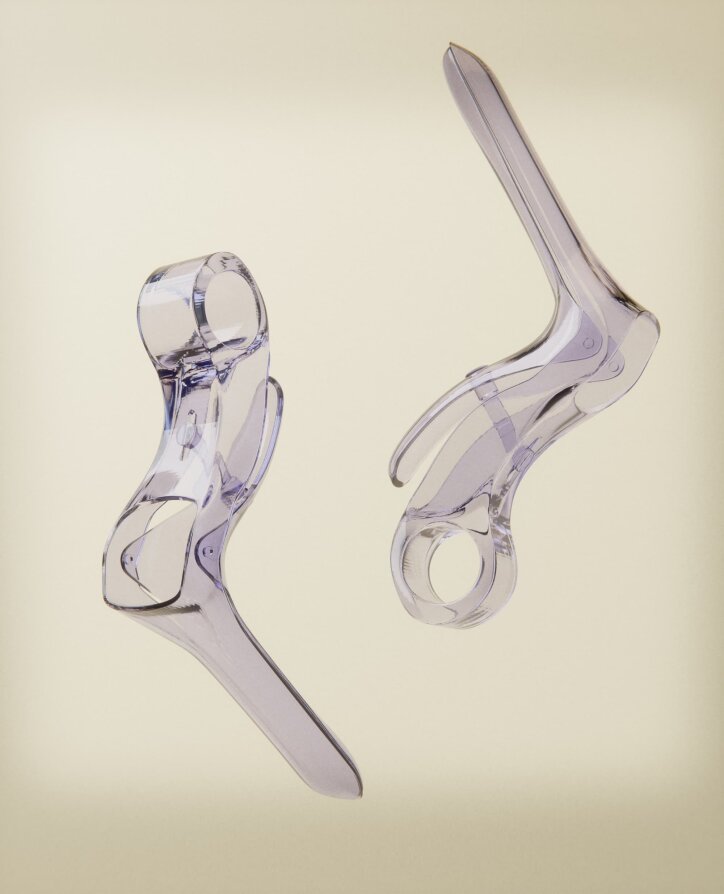 Two transparent plastic medical specula are shown against a light beige background, positioned at different angles to show their structure and details.