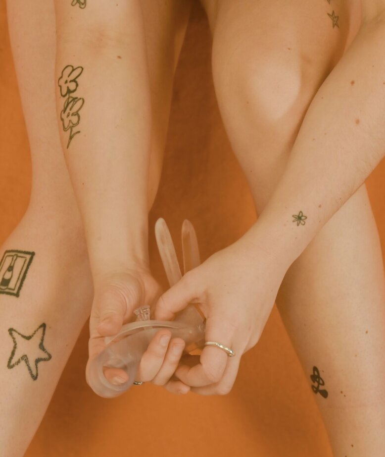 A person with various tattoos on her legs and arms is sitting in front of an orange background and holding a transparent menstrual cup. Her face is not visible in the picture.