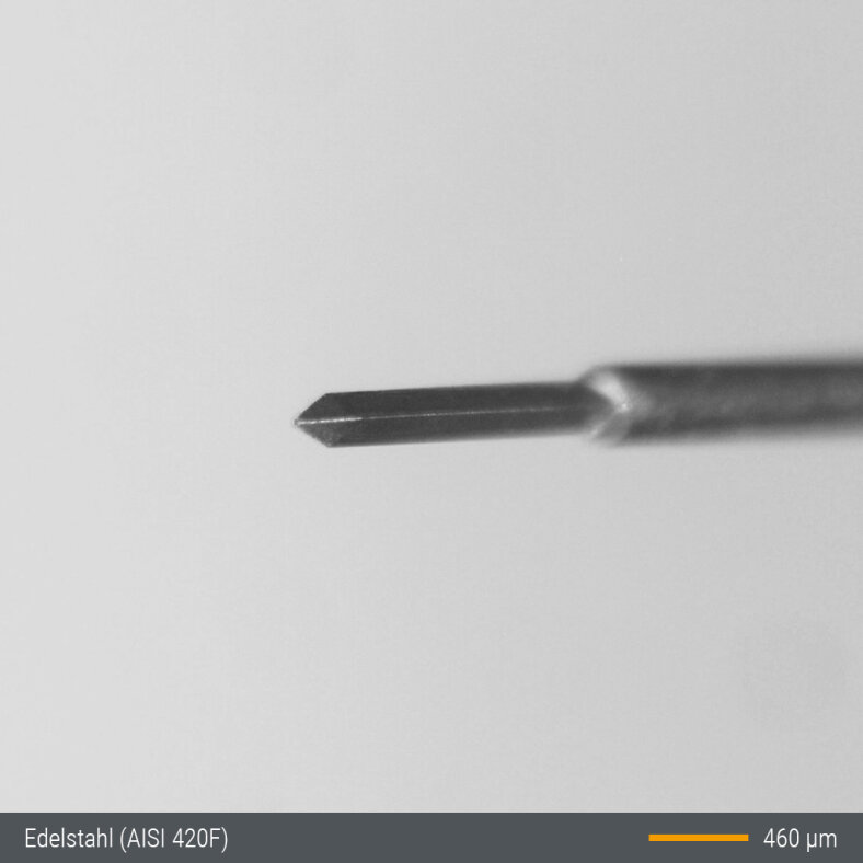 A close-up image of a fine metallic needle tip, set against a plain background, with a scale bar indicating 460 micrometers and a label noting the material as stainless steel (AISI 420F).