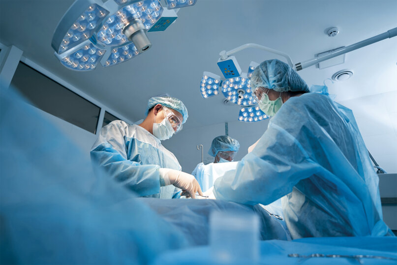Chirurgen in Kitteln, Masken und Handschuhen führen in einem modernen Operationssaal eine Operation unter hellem Operationslicht durch. Medizinische Geräte und sterile Abdeckungen umgeben das Operationsteam.