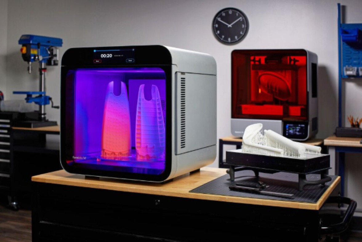Ein Desktop-3D-Drucker leuchtet violett, während er große Objekte druckt. Ein weiterer rot beleuchteter Drucker, Werkzeuge und 3D-gedruckte Modelle stehen auf Werkbänken in einer modernen Werkstatt. Im Hintergrund ist eine Wanduhr zu sehen.