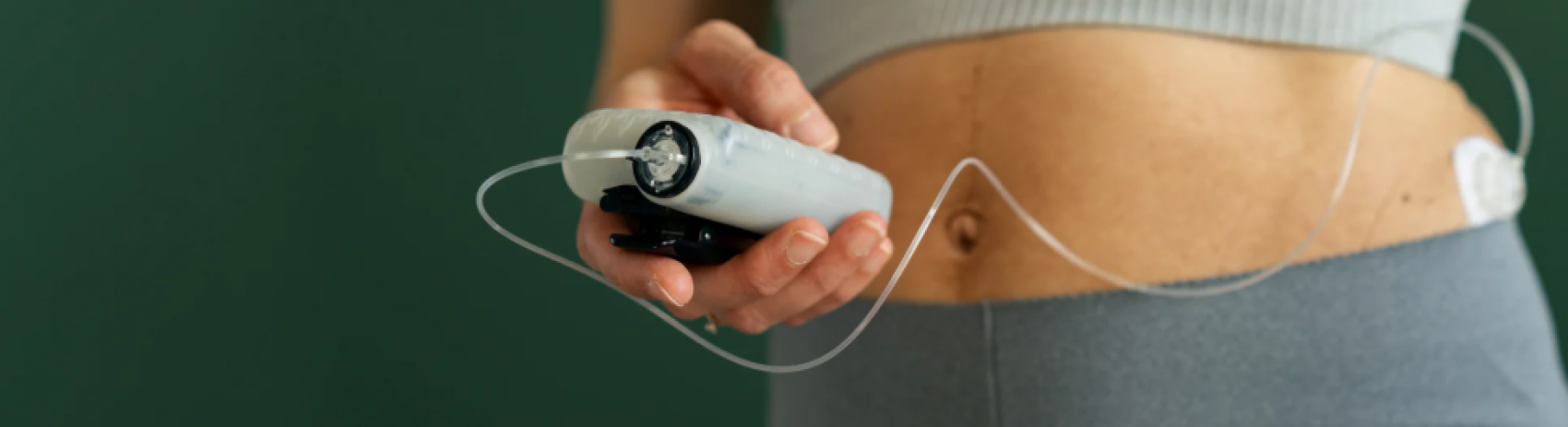 A person wearing a gray top and pants holds an insulin pump connected to their abdomen with tubing, illustrating diabetes management.