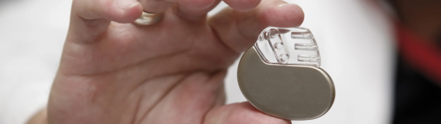 A hand holding a small medical device, likely a pacemaker, with a metallic lower half and a transparent upper section showing internal components.