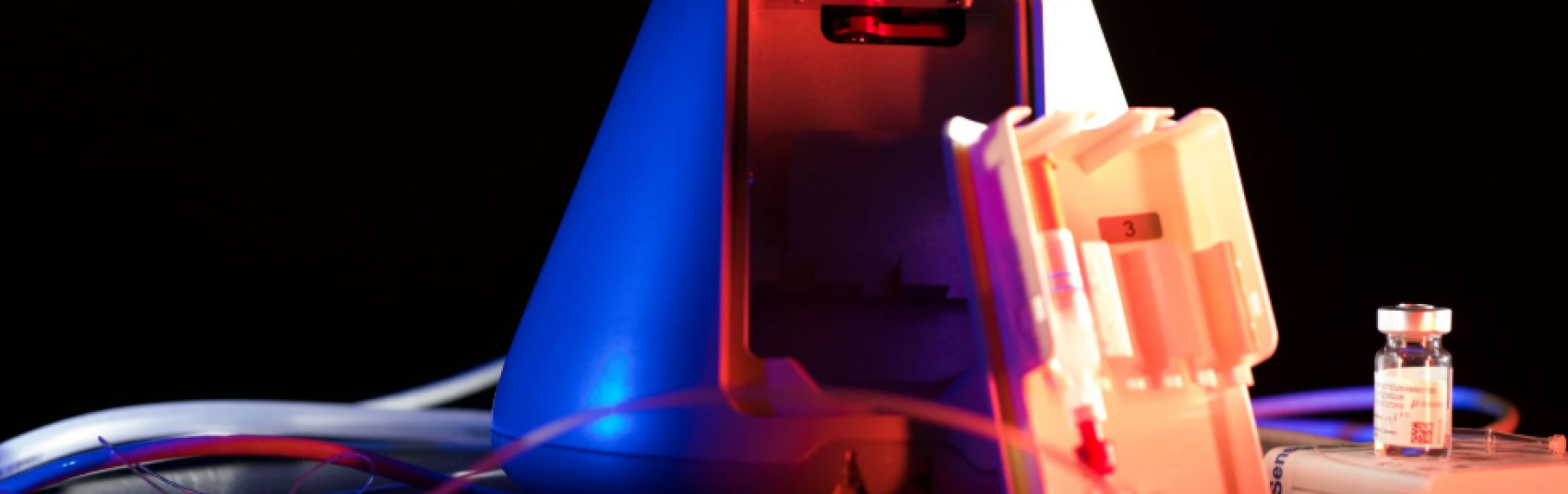 A close-up of medical equipment under blue and red lighting, including a blue device, a white plastic case with test tubes, and a small vial on a surface, with wires visible in the background.