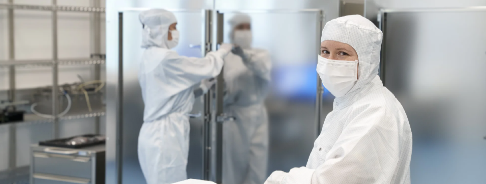Two people dressed in full white protective lab suits and masks work in a cleanroom laboratory, with one person looking at the camera and the other handling equipment near metallic cabinets.