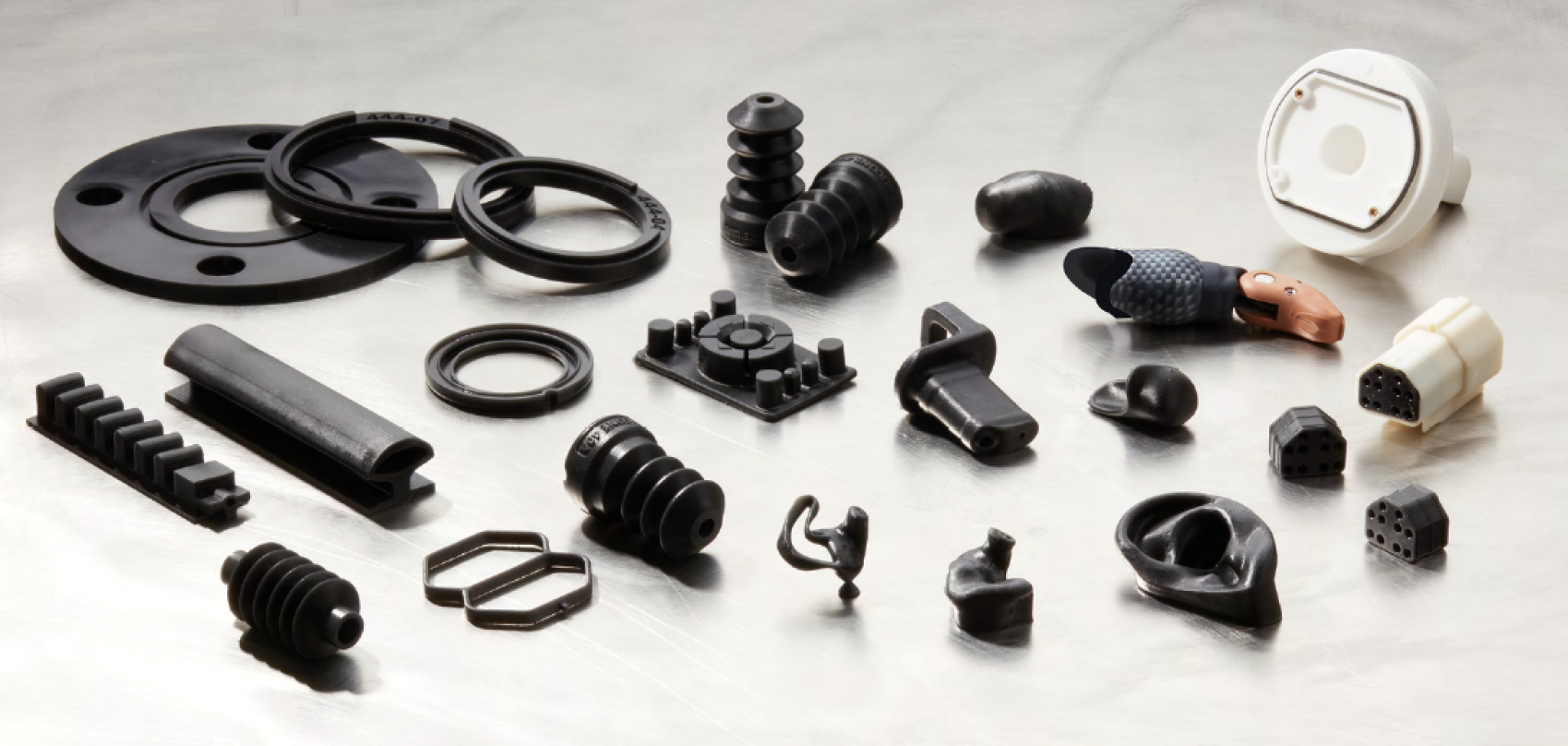 Eine Vielzahl schwarzer und weißer Formteile aus Gummi und Kunststoff aus dem Automobil- oder Industriebereich, darunter Dichtungen, Stecker und Verbinder, sind auf einer reflektierenden Oberfläche angeordnet.