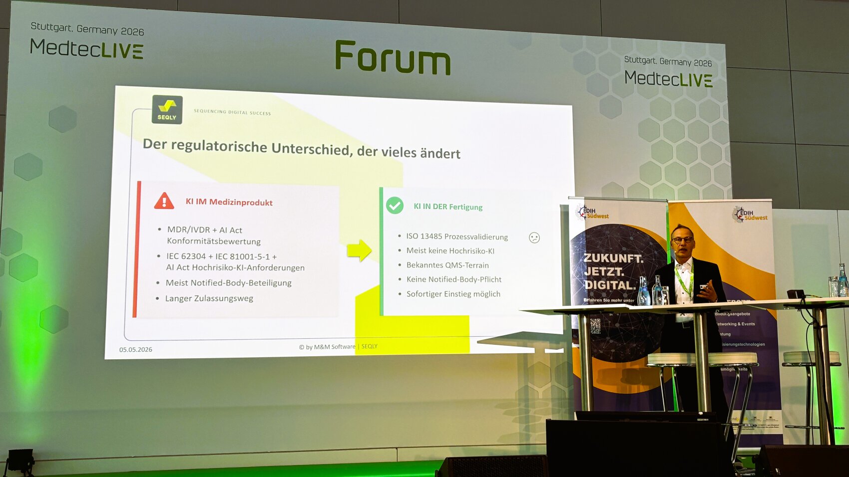 Ein Referent steht auf der Bühne der MedtecLIVE 2026 in Stuttgart am Rednerpult. Hinter ihm ist auf einer Leinwand eine deutschsprachige Folie zu sehen, auf der die regulatorischen Anforderungen an Medizinprodukte im Rahmen einer Konferenz verglichen werden.