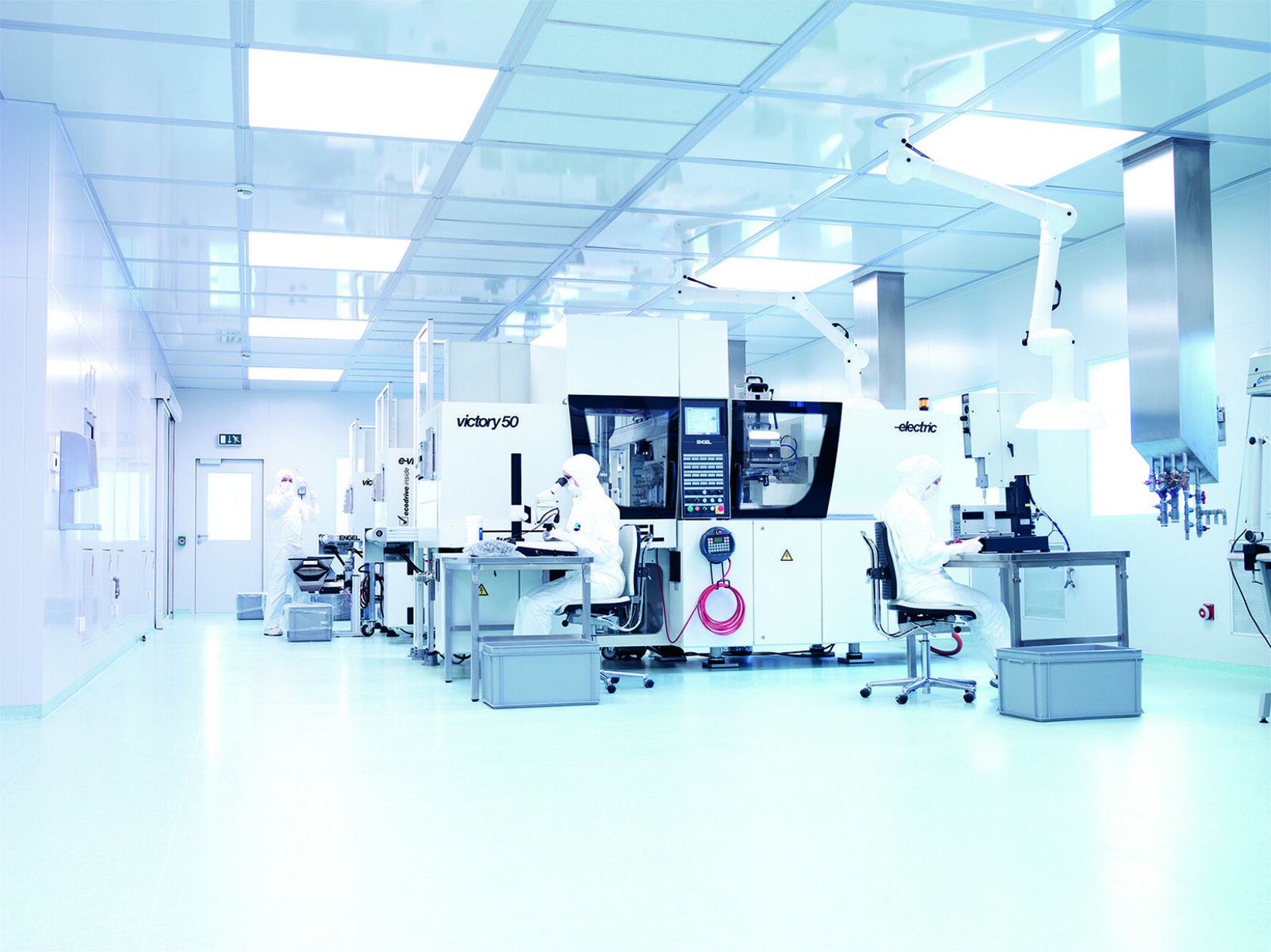 Wissenschaftler in Ganzkörper-Schutzanzügen arbeiten in einem hellen, sauberen Labor mit weißen Wänden und Böden, mit Maschinen, Oberlichtern und geordneten Arbeitsplätzen an modernen Geräten.